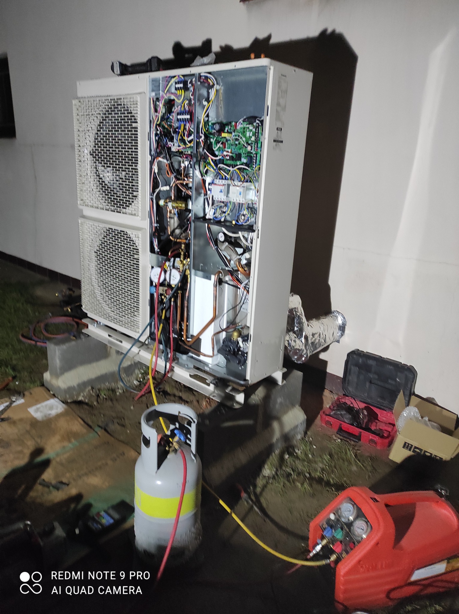 Otwarta obudowa zewnętrznej jednostki klimatyzacji typu split z widocznymi podzespołami elektrycznymi i rurami miedzianymi, podłączona do butli z czynnikiem chłodniczym i czerwonej stacji odzysku...