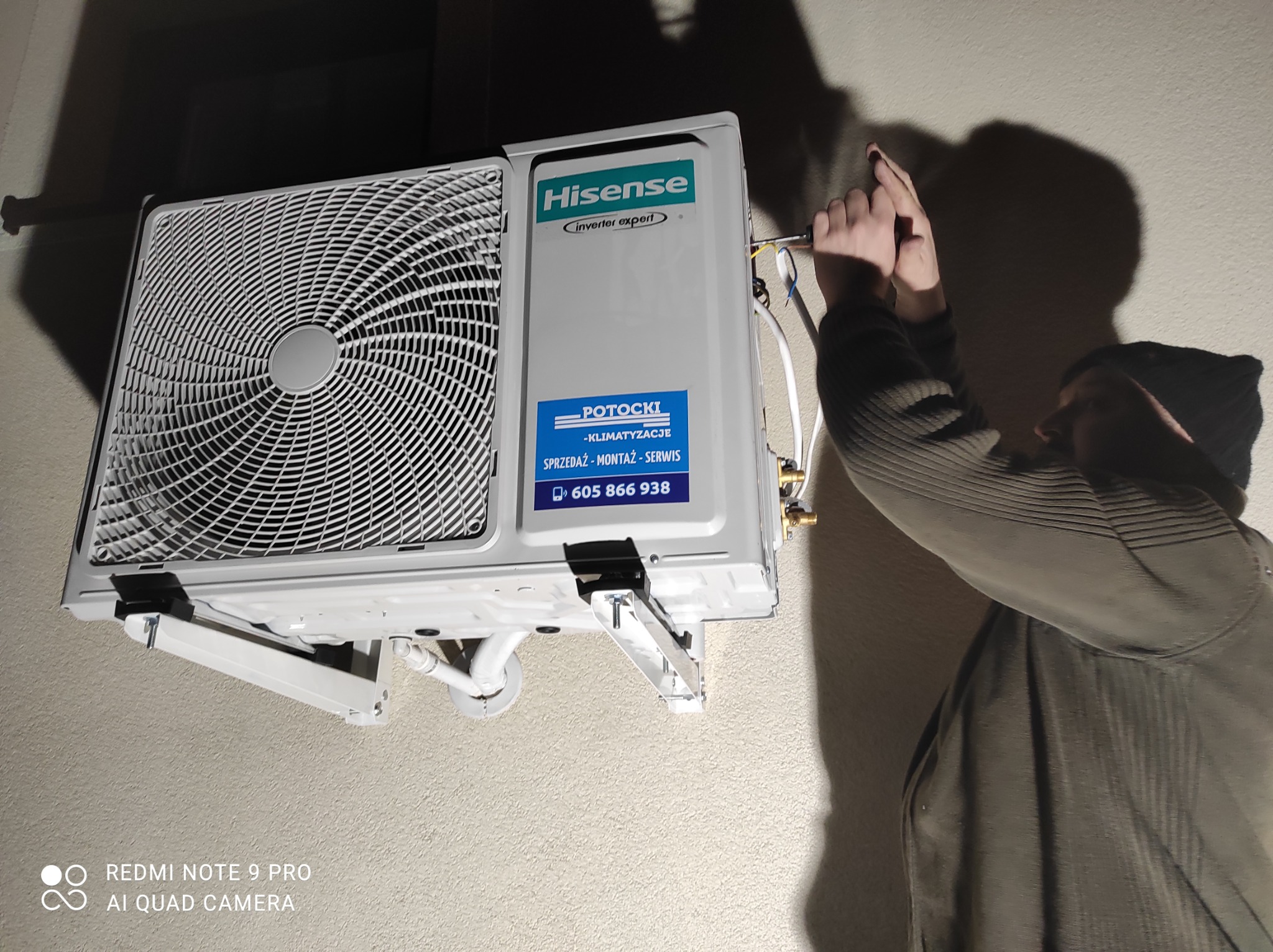 Instalacja klimatyzatora Hisense na ścianie. Widoczny monter w trakcie podłączania przewodów elektrycznych. Na urządzeniu naklejka z nazwą firmy Potocki Klimatyzacje i numerem telefonu.