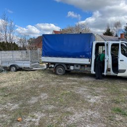 OLBUD sp. z o.o. - Biały samochód dostawczy z niebieską plandeką i przyczepką na trawiastym terenie, pracownik w zielonej koszulce wsiada do kabiny.