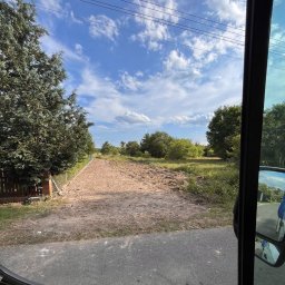 OLBUD sp. z o.o. - Wyrównany teren pod budowę, widok zza szyby pojazdu. Przygotowany grunt z widocznymi śladami prac ziemnych, otoczony zielenią i ogrodzeniem. Błękitne niebo z chmurami w tle.