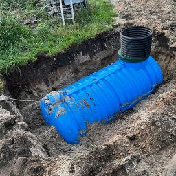 OLBUD sp. z o.o. - Niebieski, plastikowy zbiornik zakopany w ziemi, z widocznym czarnym kominem. W tle trawa, drabina i fragment samochodu. Instalacja szamba.