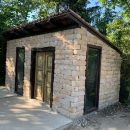 Niewielka budowla z piaskowych bloków z ciemnymi ramami okiennymi i drzwiami w ogrodzie, widoczne elementy drenażu liniowego na betonowej wylewce.