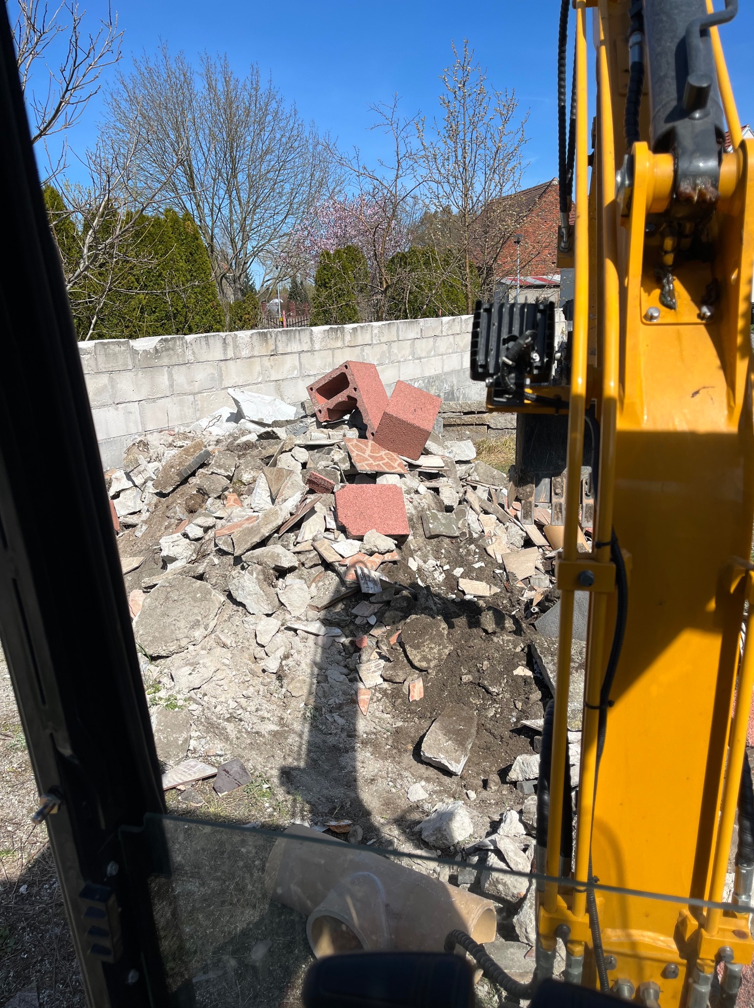 Widok z kabiny minikoparki na stertę gruzu i ziemi z fragmentami cegieł, z betonowym murem w tle i drzewami na horyzoncie w słoneczny dzień.
