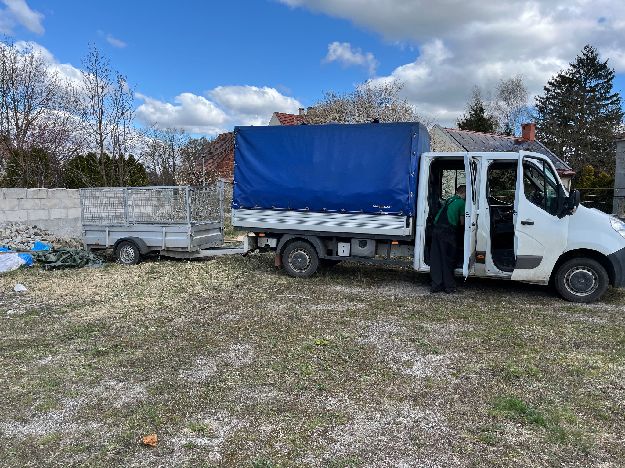 Biały samochód dostawczy z niebieską plandeką i przyczepką na trawiastym terenie, pracownik w zielonej koszulce wsiada do kabiny.