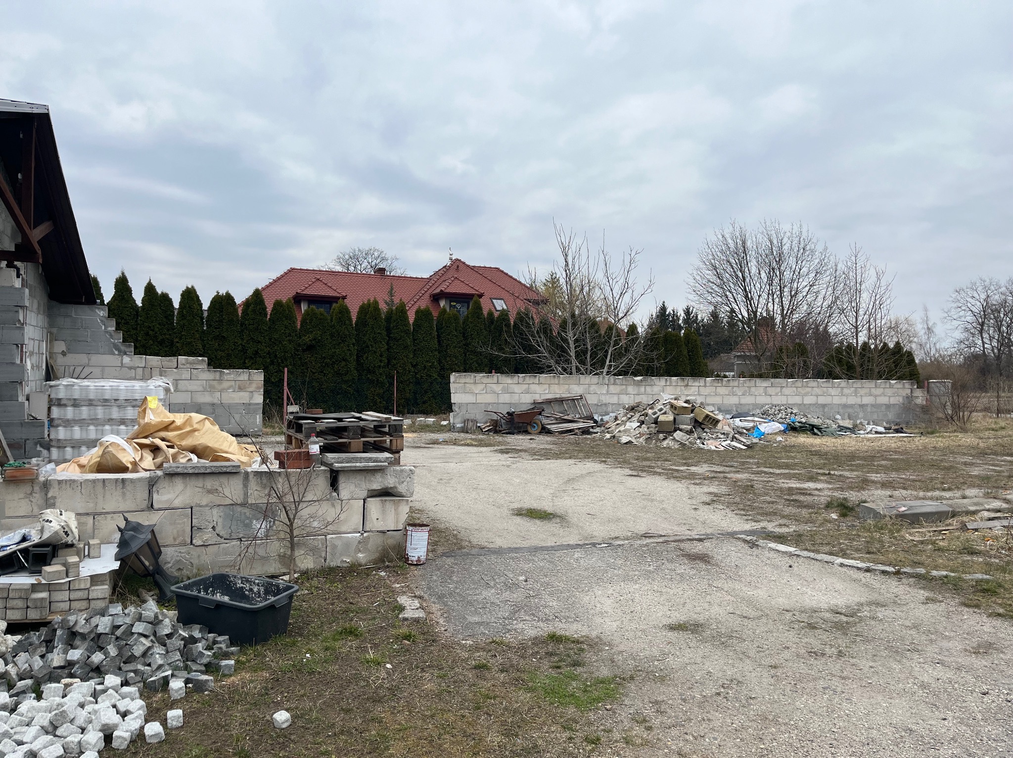 Niedokończony mur z betonowych bloczków na posesji z widocznymi stertami gruzu i resztek materiałów budowlanych, w tle rząd tuj i dom z czerwoną dachówką.