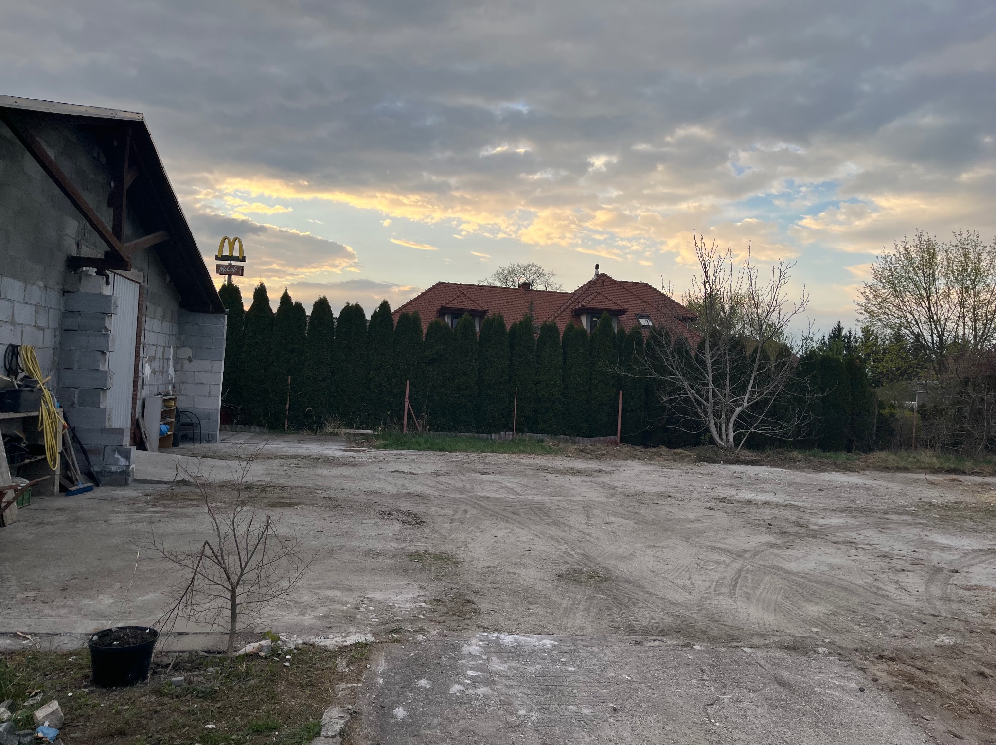 Przygotowany teren pod budowę z widocznymi śladami opon, budynkiem gospodarczym z bloczków betonowych, wysokimi tujami w tle i znakiem McDonald's w oddali.