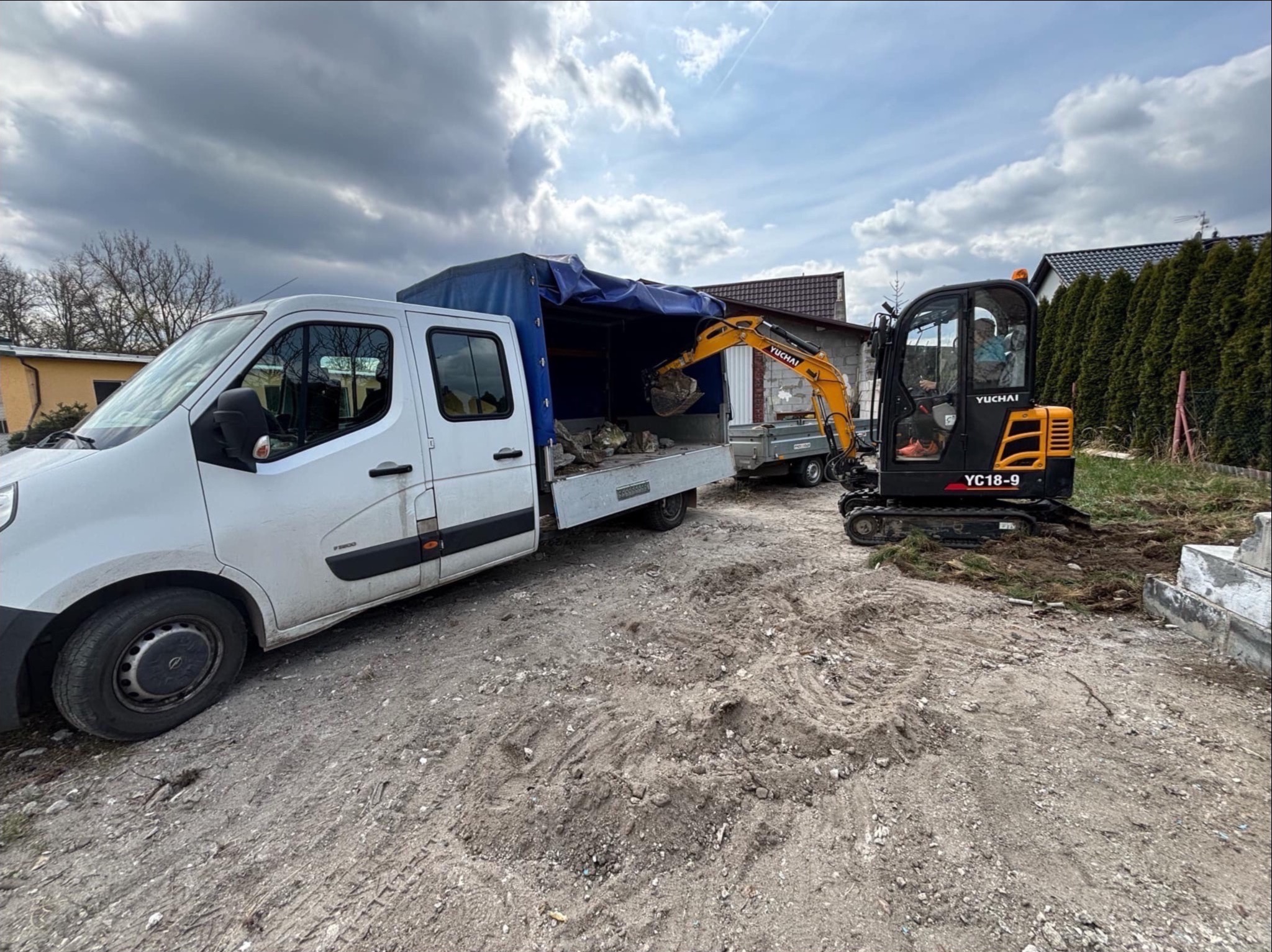 Żółto-czarna minikoparka Yuchai YC18-9 ładuje gruz do skrzyni białego, zakrytego plandeką samochodu dostawczego na terenie budowy.