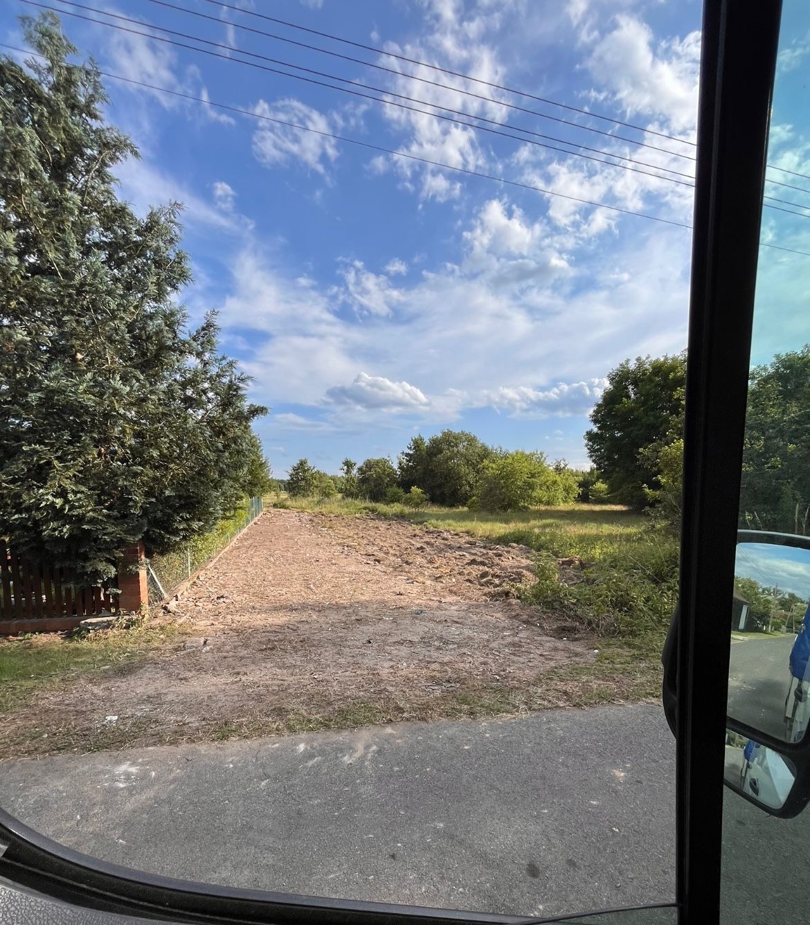 Wyrównany teren pod budowę, widok zza szyby pojazdu. Przygotowany grunt z widocznymi śladami prac ziemnych, otoczony zielenią i ogrodzeniem. Błękitne niebo z chmurami w tle.