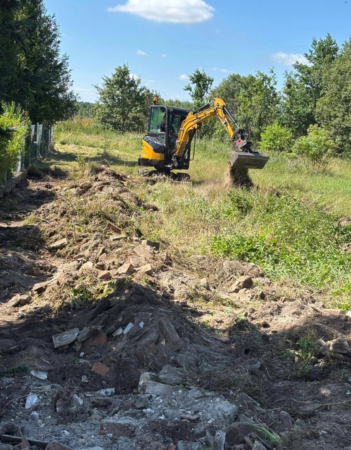 Żółta minikoparka w trakcie prac ziemnych na zarośniętym terenie. W tle drzewa i niebo. Na pierwszym planie gruz i ziemia. Widoczne ślady pracy maszyny.