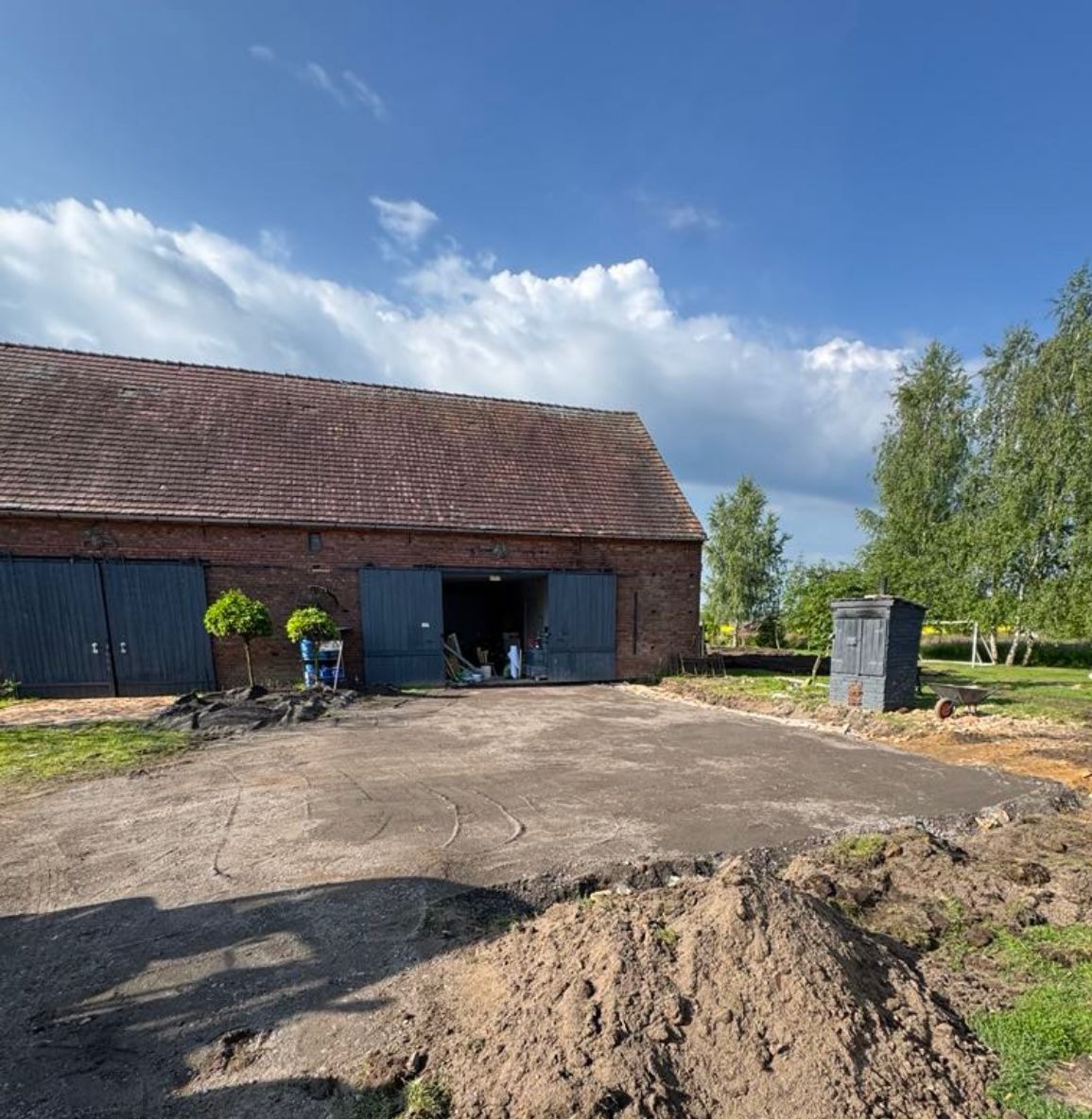 Przygotowany teren pod budowę z widocznym budynkiem gospodarczym z czerwonej cegły i otwartymi wrotami. W tle drzewa i błękitne niebo z chmurami.
