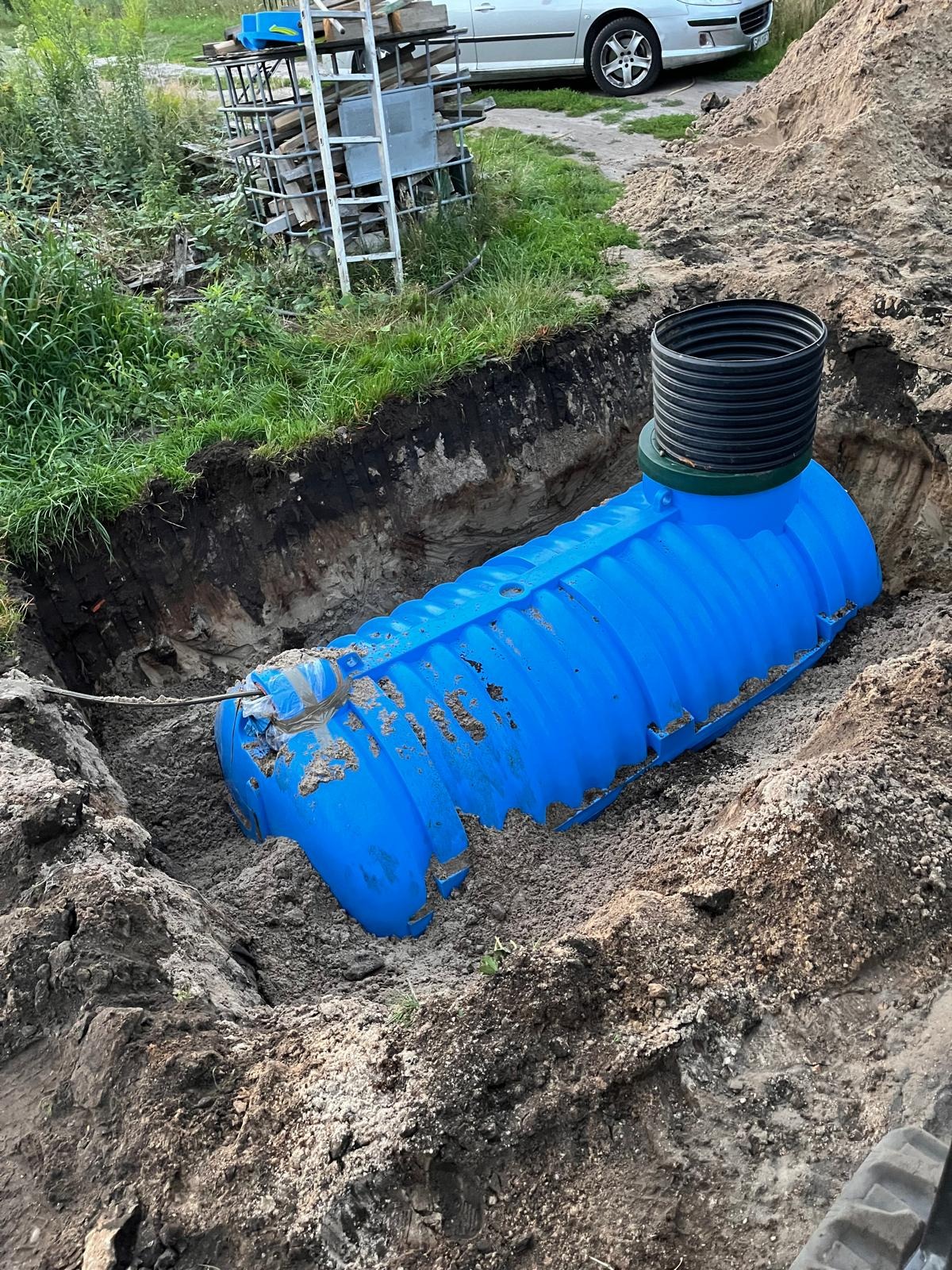 Niebieski, plastikowy zbiornik zakopany w ziemi, z widocznym czarnym kominem. W tle trawa, drabina i fragment samochodu. Instalacja szamba.