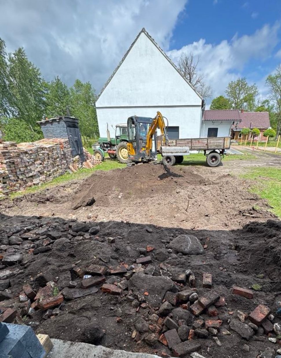 Żółta minikoparka z przyczepą na wykopie w Dębince. W tle budynek i sterta cegieł. Prace ziemne w toku, widoczne ślady po maszynie.