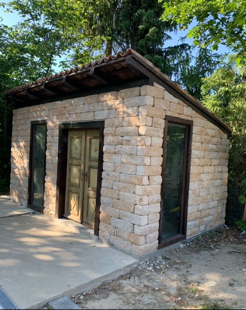 Niewielka budowla z piaskowych bloków z ciemnymi ramami okiennymi i drzwiami w ogrodzie, widoczne elementy drenażu liniowego na betonowej wylewce.