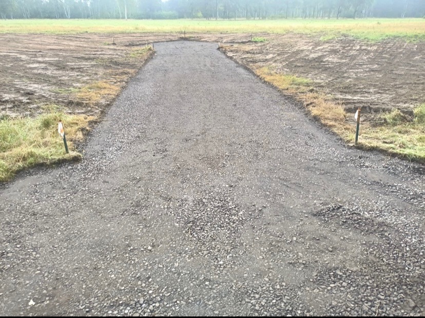 Świeżo wysypana żwirowa droga dojazdowa na terenie budowy, wyznaczona palikami, z widocznym przygotowanym terenem pod dalsze prace.