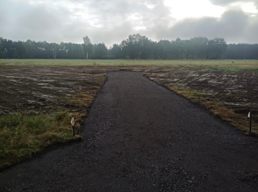Przygotowany teren pod budowę z wyrównaną drogą dojazdową z kruszywa; widoczne znaczniki geodezyjne i ślady po maszynach budowlanych na wilgotnej glebie, w tle zalesiony horyzont pod pochmurnym...