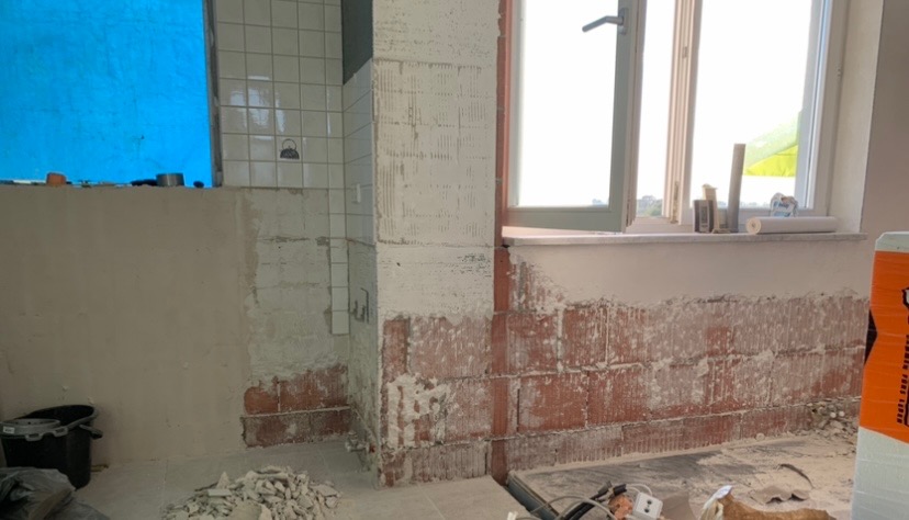 Wnętrze łazienki w trakcie remontu: częściowo położone płytki, odsłonięte cegły, resztki gruzu na podłodze, okno z widokiem na zewnątrz, zabezpieczone okno folią. Widoczny etap przygotowania ścian...