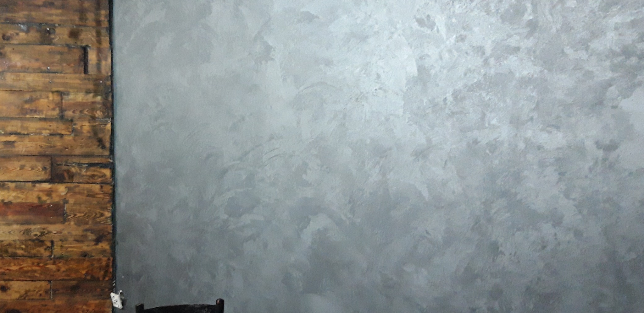 Dekoracyjna ściana w odcieniach szarości z efektem perłowym, kontrastująca z fragmentem ściany wyłożonej starymi, ciemnymi deskami.