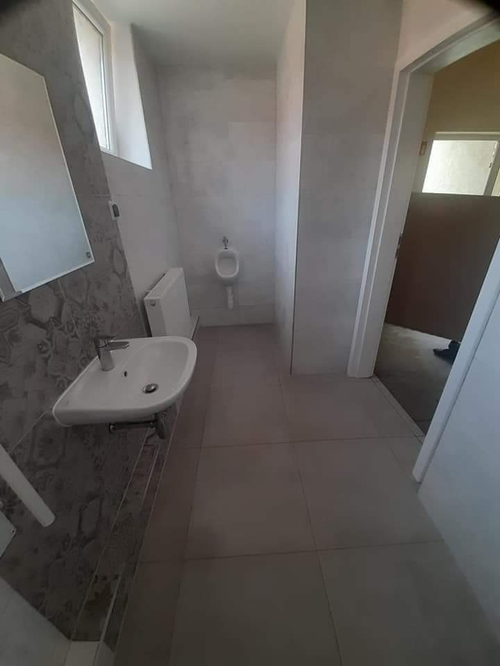 Wykończona łazienka z umywalką, grzejnikiem i pisuarzem w minimalistycznym stylu, szare płytki na podłodze i wzorzyste na ścianie przy umywalce, widoczny fragment otwartych drzwi.