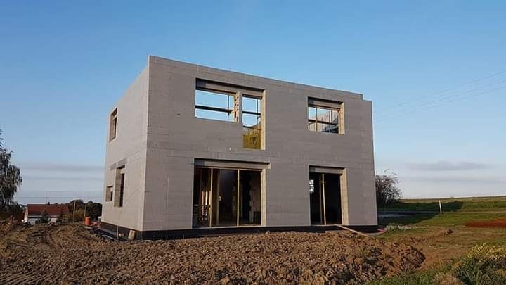 Dwukondygnacyjny dom w stanie surowym otwartym z szarych bloczków, widoczne otwory okienne i drzwiowe, teren wokół przygotowany pod dalsze prace.