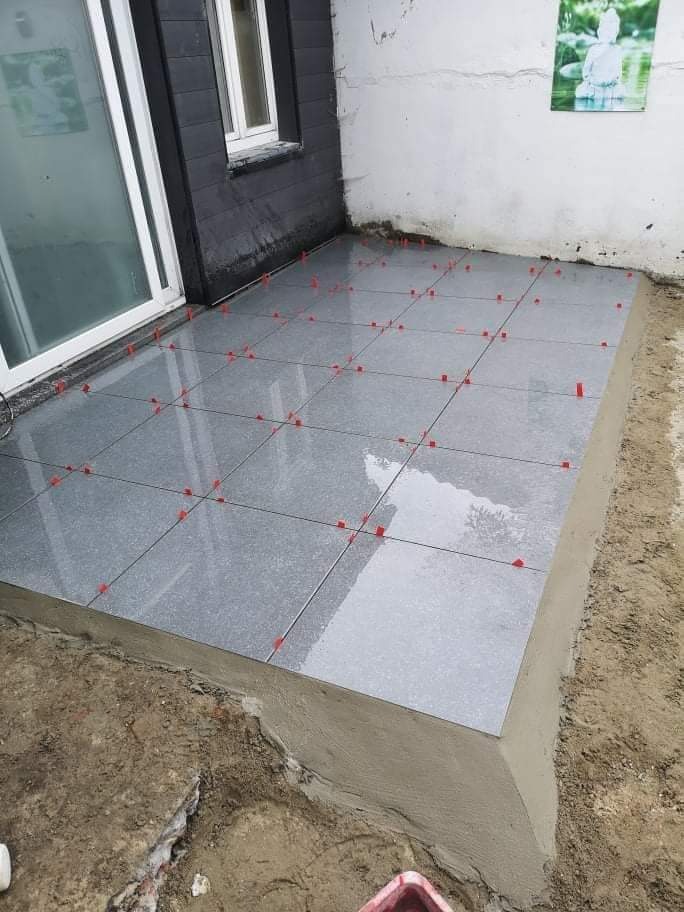 Świeżo ułożone, błyszczące, szare płytki na tarasie, z czerwonymi klipsami poziomującymi, widoczne niedoskonałości podłoża i ślady po pracach budowlanych.