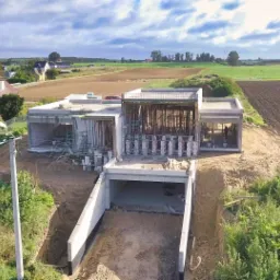 Widok z lotu ptaka na budowę domu z betonowym fundamentem i wjazdem do garażu podziemnego, otoczonego krajobrazem wiejskim z polami i zabudowaniami w oddali, w słoneczny dzień.