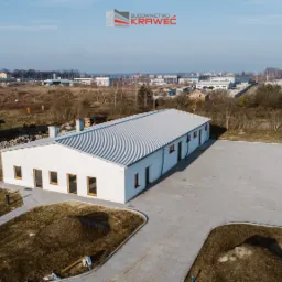 Zdjęcie z lotu ptaka przedstawia ukończony, parterowy budynek o białych ścianach i szarym, blaszanym dachu, z utwardzonym placem przed wejściem, w tle zabudowa jednorodzinna i logo firmy budowlanej.