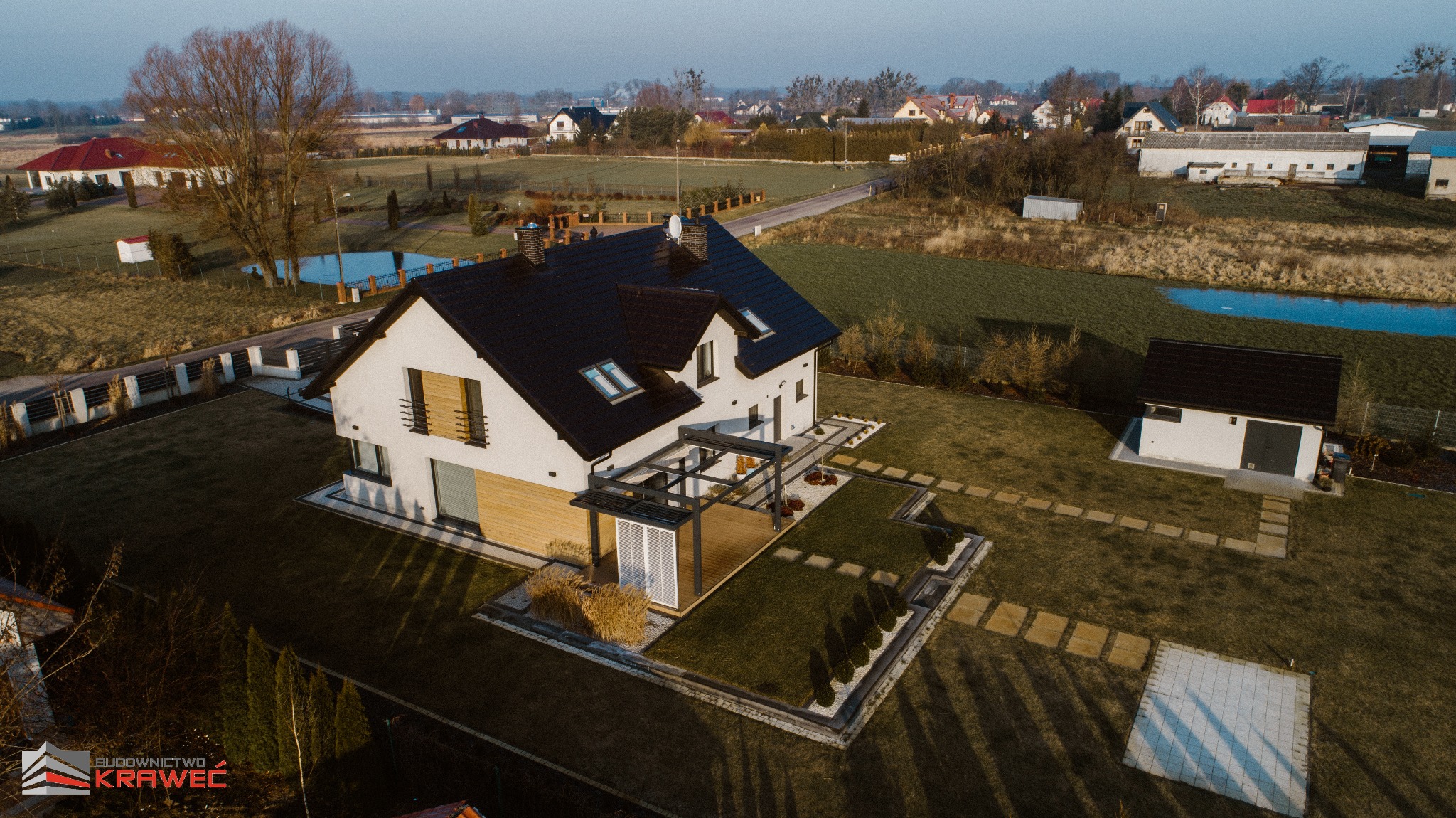 Nowoczesny dom jednorodzinny z czarnym dachem i elementami drewnianymi, widok z drona na zadbany ogród z pergolą i ścieżkami z płyt.