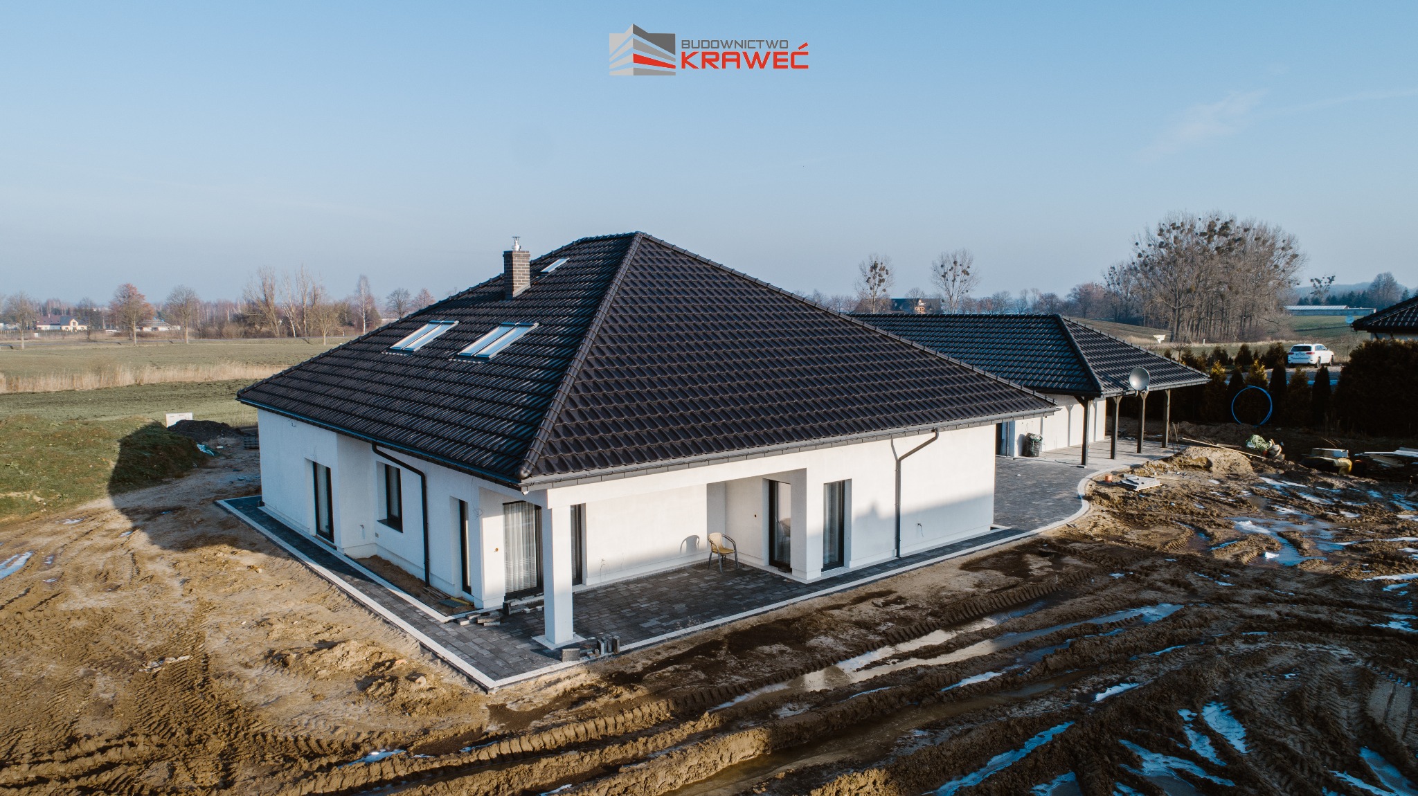 Nowoczesny dom jednorodzinny z białymi ścianami i ciemną dachówką, widok z góry na posesję w trakcie zagospodarowywania terenu, z widocznymi śladami opon w błocie i logo firmy budowlanej.
