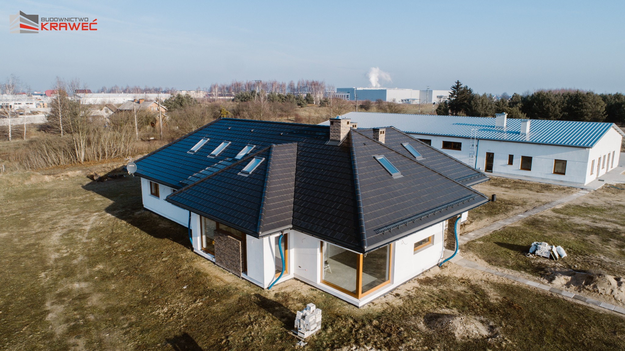 Nowoczesny, parterowy dom z ciemną dachówką, dużymi oknami i wykończeniem z kamienia elewacyjnego, widziany z lotu ptaka, z widocznym logo firmy budowlanej w lewym górnym rogu.