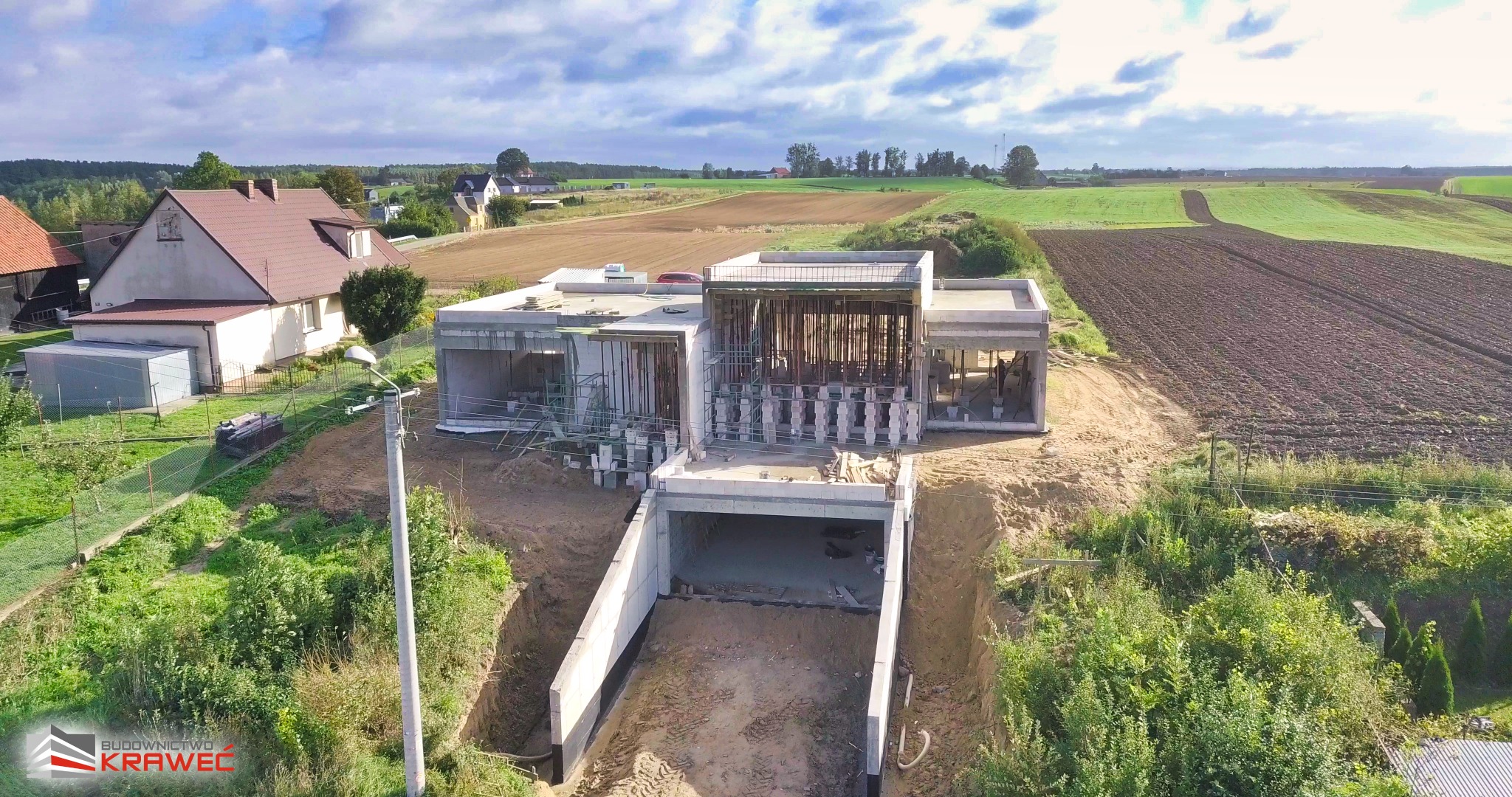 Widok z lotu ptaka na budowę domu z betonowym fundamentem i wjazdem do garażu podziemnego, otoczonego krajobrazem wiejskim z polami i zabudowaniami w oddali, w słoneczny dzień.