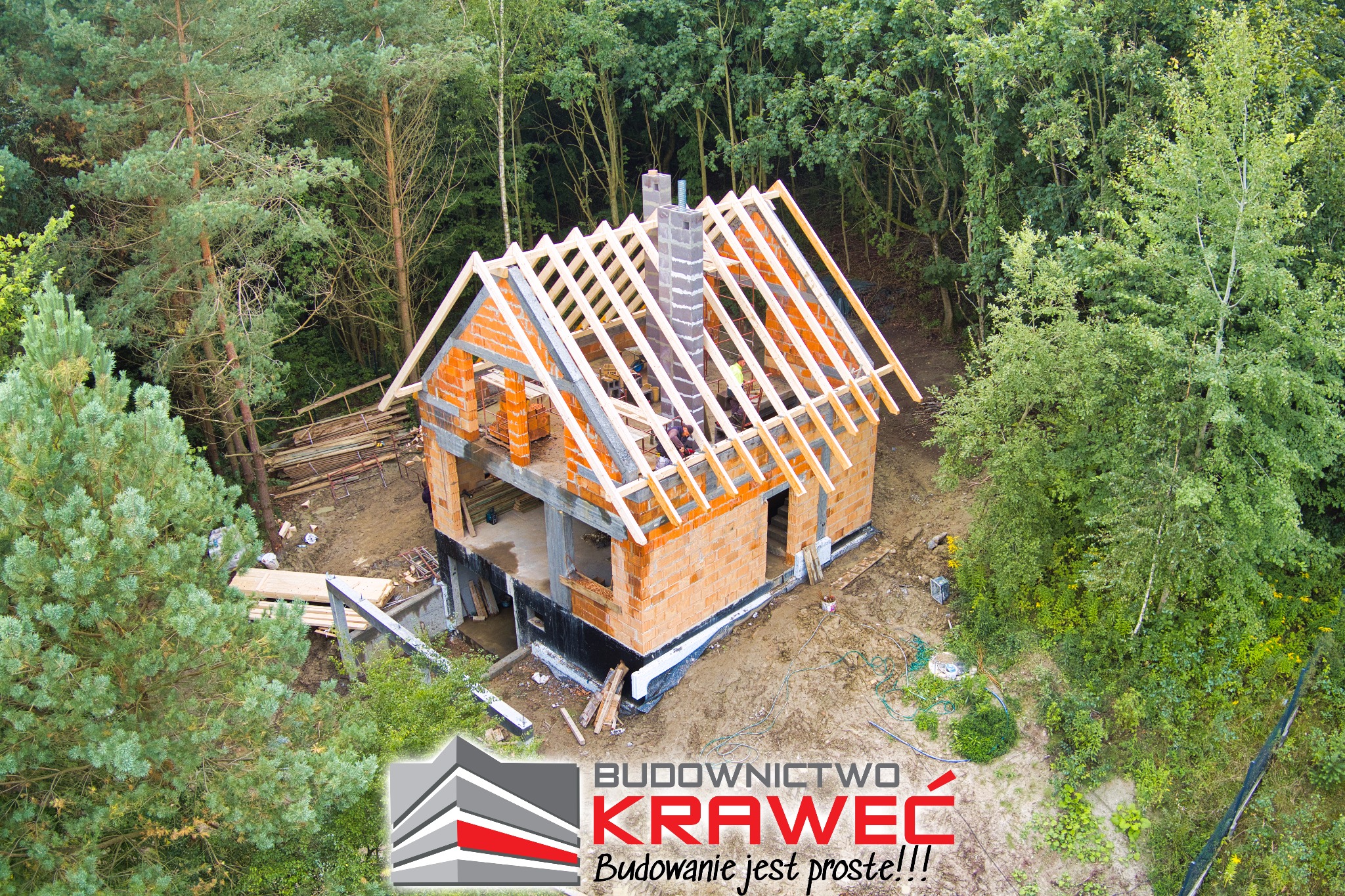 Zdjęcie z lotu ptaka przedstawia budowę domu z czerwonej cegły z drewnianą konstrukcją dachu w otoczeniu drzew, z widocznym logo firmy budowlanej w rogu.