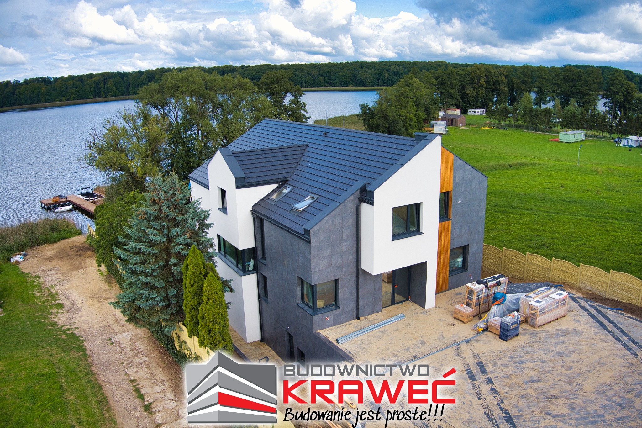 Nowoczesny dom z ciemną dachówką i elewacją w odcieniach szarości i bieli, widok z lotu ptaka na tle jeziora i zieleni, częściowo wykończony teren wokół domu z paletami materiałów budowlanych.