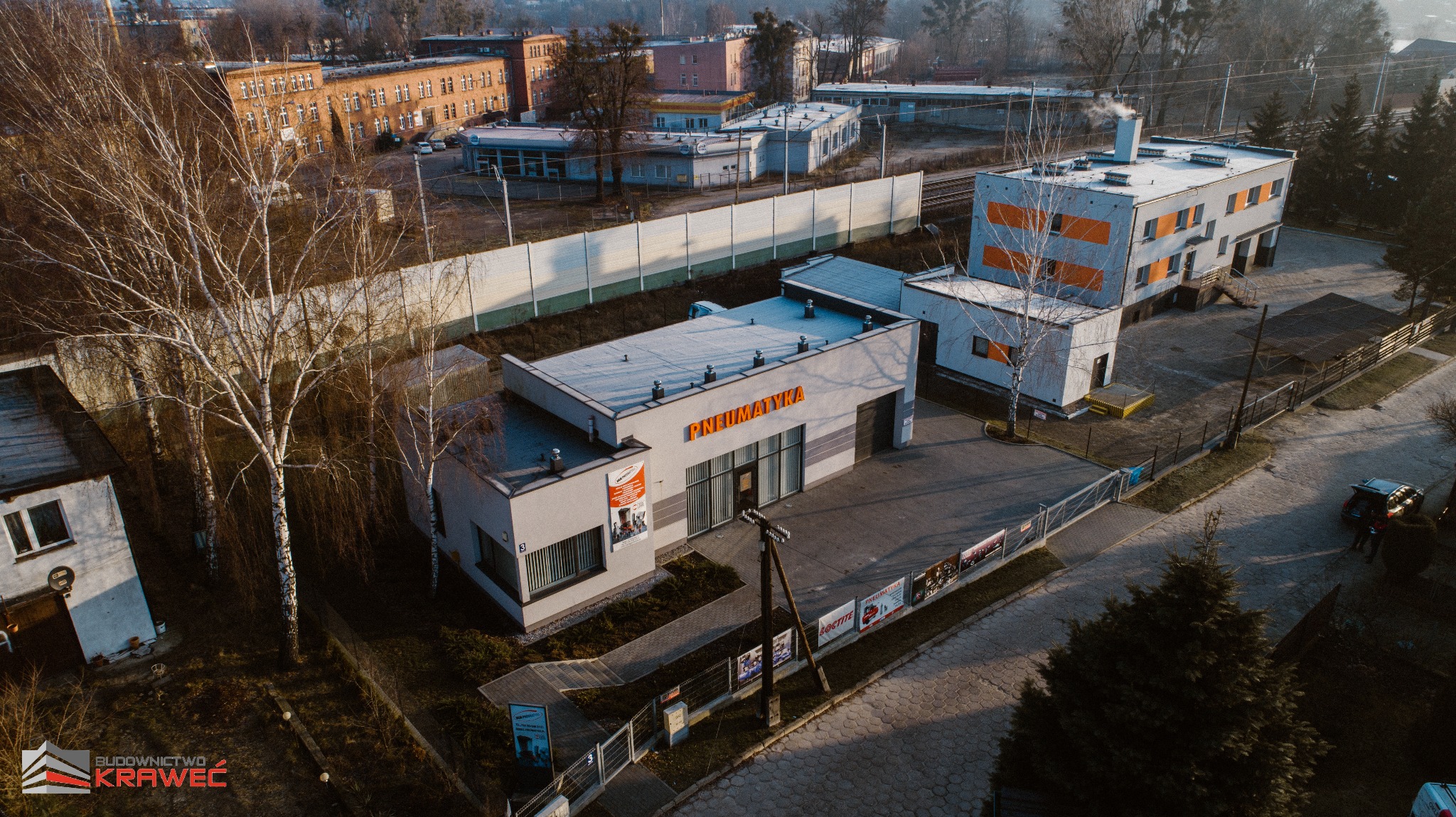Widok z góry na budynek firmy 'Pneumatyka' z szarymi ścianami i pomarańczowym napisem, otoczony drzewami i innymi budynkami w tle, uchwycony w porannym świetle.