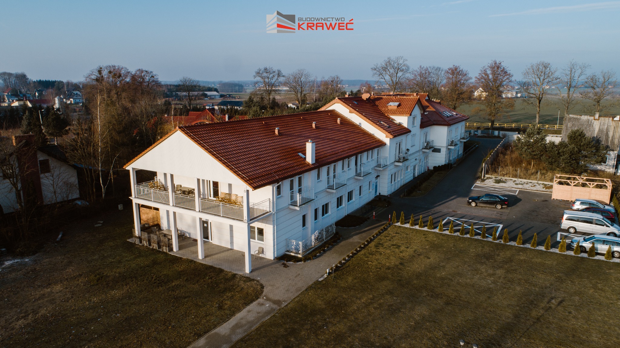 Widok z lotu ptaka na dwukondygnacyjny budynek z białymi ścianami i czerwoną dachówką, z balkonami i parkingiem z kilkoma samochodami, otoczony zielenią i innymi budynkami w tle, z logo firmy...