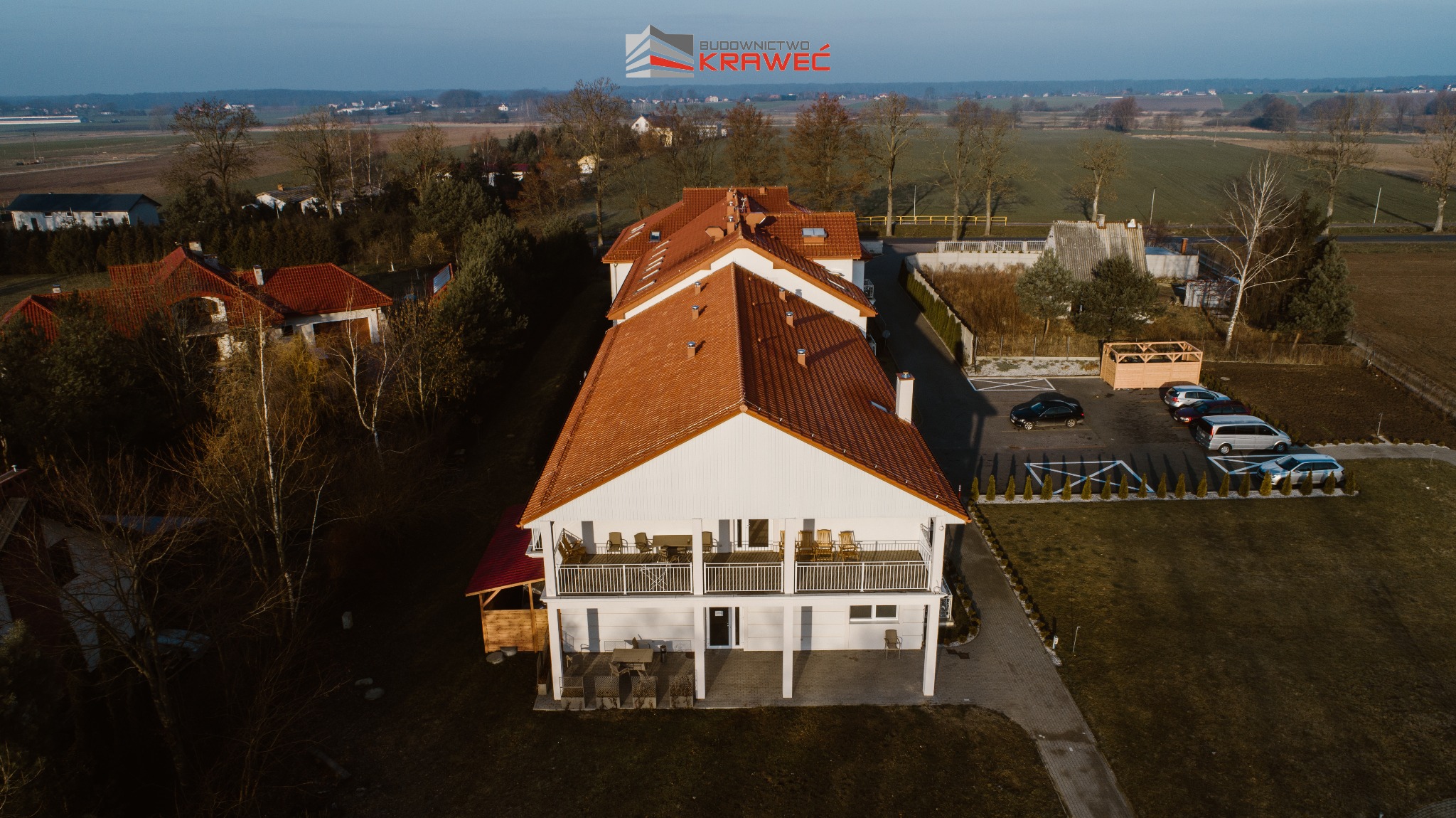 Widok z drona na dwukondygnacyjny budynek z czerwonym dachem i białymi ścianami, z balkonami wyposażonymi w meble ogrodowe, otoczony zielenią i parkingiem z samochodami.