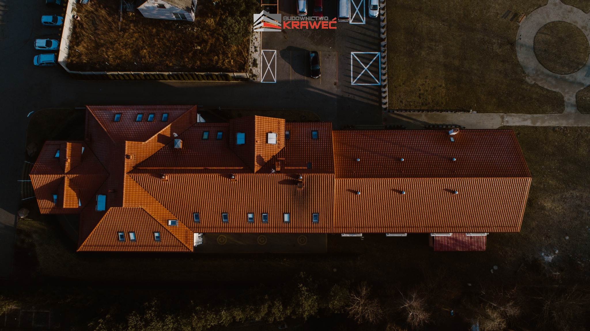 Widok z góry na duży budynek z czerwoną dachówką, parkingiem z samochodami i trawnikiem, w tle widoczne drzewa i logo firmy budowlanej.