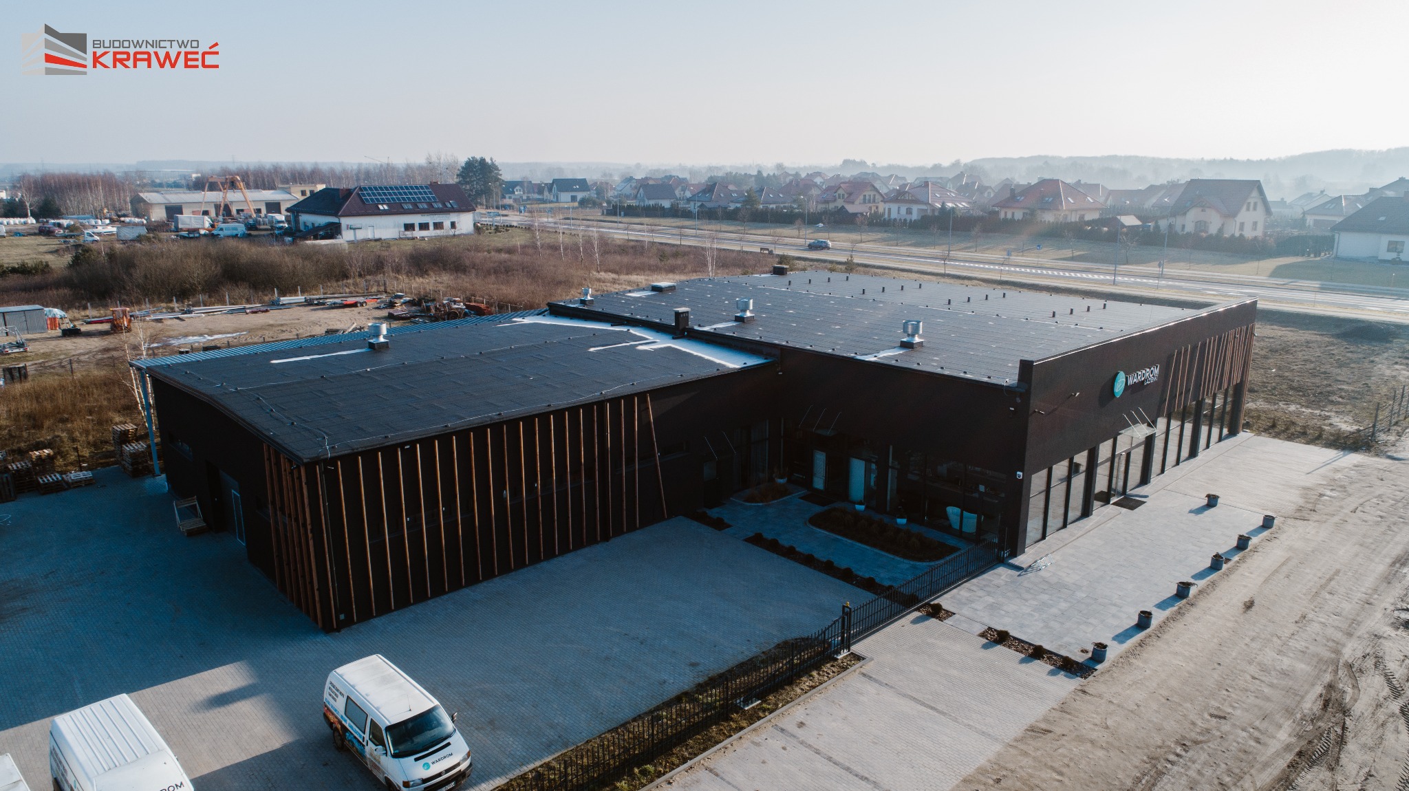 Zdjęcie z lotu ptaka przedstawia ukończony budynek biurowy z ciemną elewacją i pionowymi drewnianymi elementami dekoracyjnymi, z logo 'WARDROM' na fasadzie, z dwoma samochodami dostawczymi...