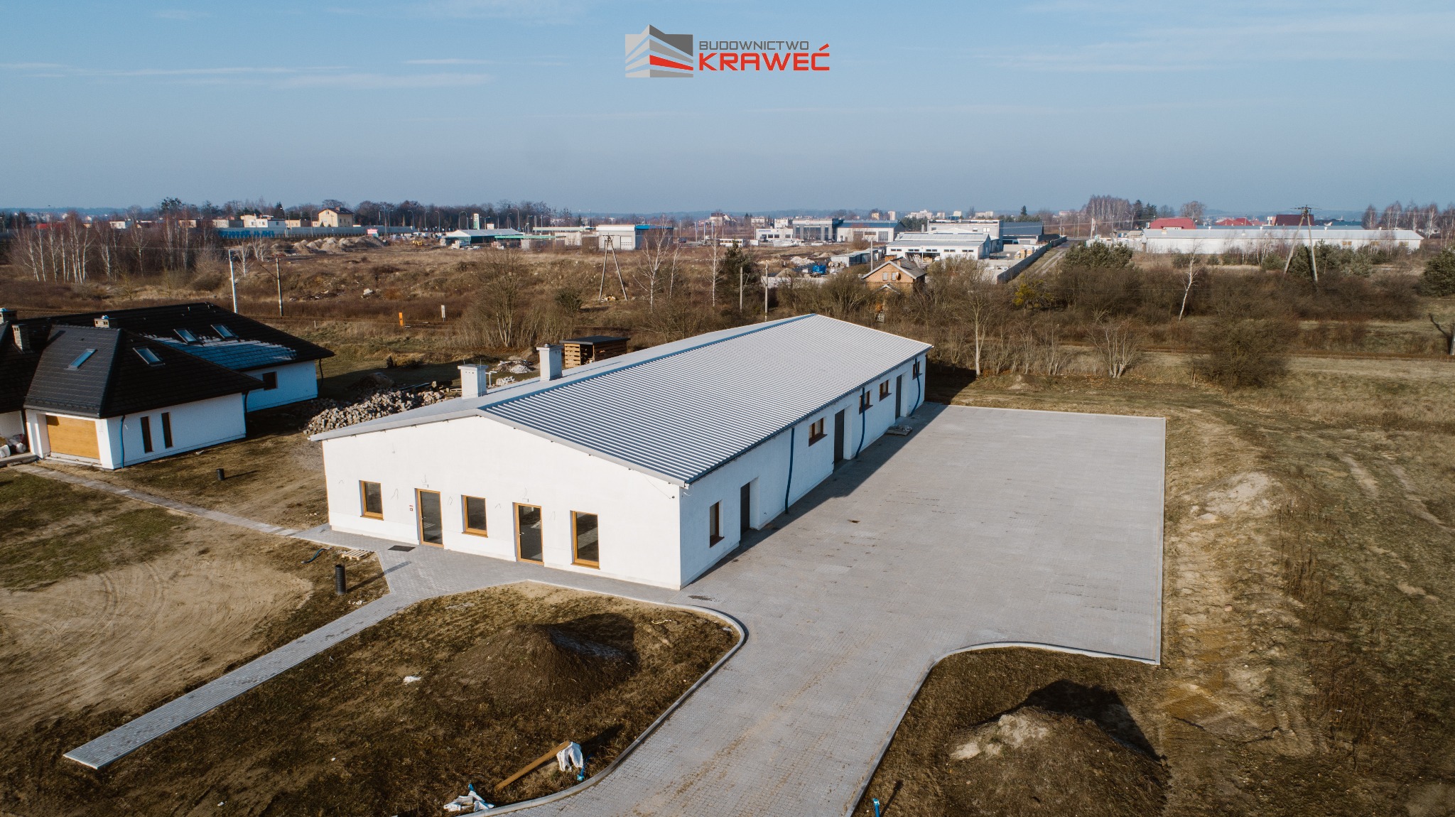 Zdjęcie z lotu ptaka przedstawia ukończony, parterowy budynek o białych ścianach i szarym, blaszanym dachu, z utwardzonym placem przed wejściem, w tle zabudowa jednorodzinna i logo firmy budowlanej.