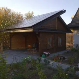 Drewniana altana z panelami słonecznymi na dachu, widok z ogrodu z kwiatami i ścieżką z kostki brukowej.