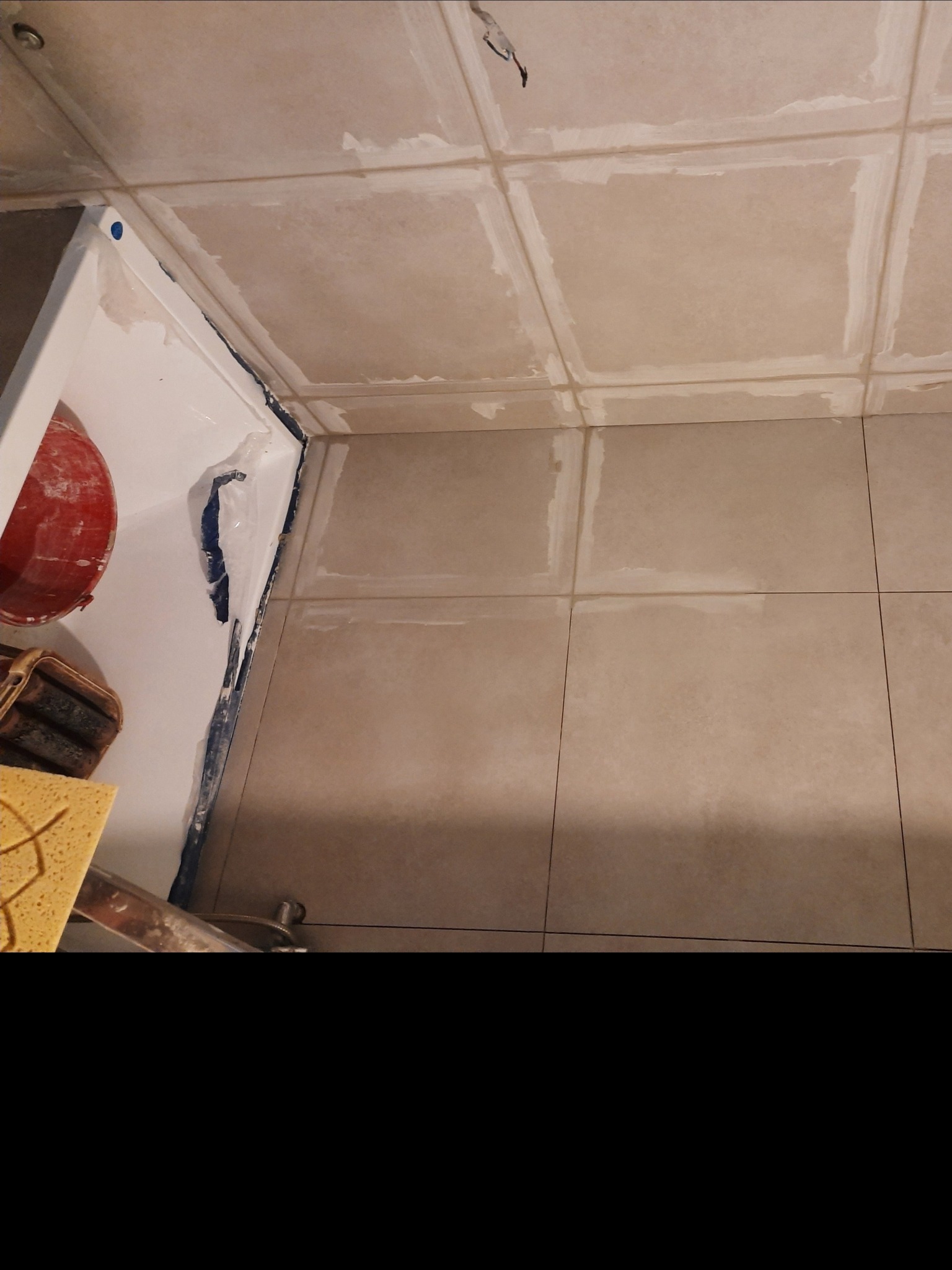 Przygotowanie do fugowania płytek w łazience: widoczna wanna z zabezpieczeniem, beżowe płytki podłogowe i sufitowe z wypełnionymi fugami, czerwone wiadro oraz gąbki.