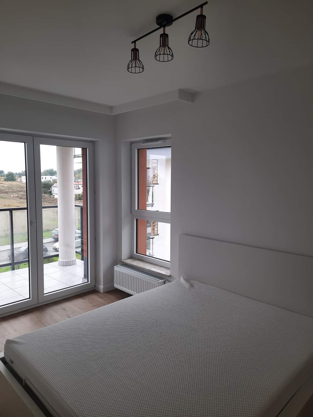 Jasne wnętrze sypialni z białymi ścianami, widokiem na balkon i parkietem, oświetlone trzema lampami w stylu industrialnym