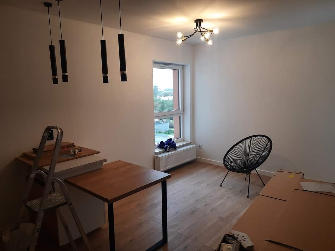 Pokój po remoncie: świeżo pomalowane ściany w odcieniach bieli, drewniana podłoga, nowoczesne lampy sufitowe, czarne krzesło plecione, drabina malarska i meble zabezpieczone kartonami.
