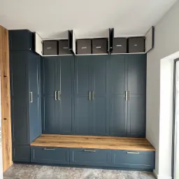 Granatowa zabudowa meblowa z drewnianym siedziskiem i szafkami do sufitu, niektóre z nich otwarte, ukazujące szare pojemniki.