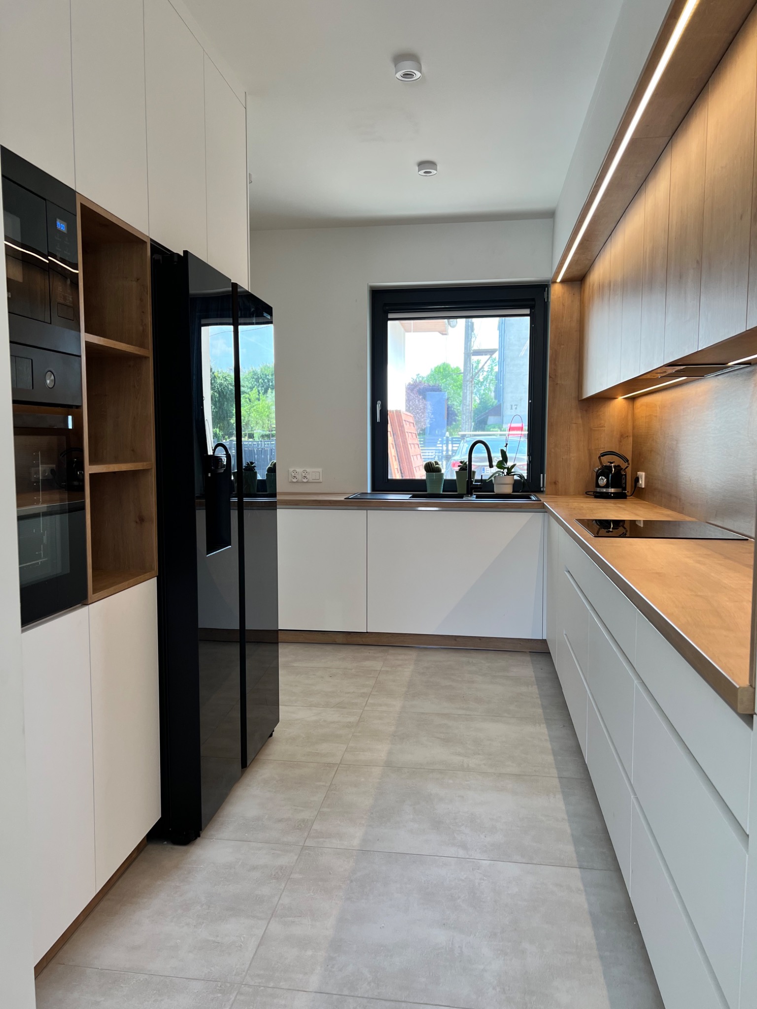 Nowoczesna kuchnia z białymi frontami szafek, drewnianymi blatami i oświetleniem LED, czarną lodówką side-by-side i widokiem na zieleń za oknem.