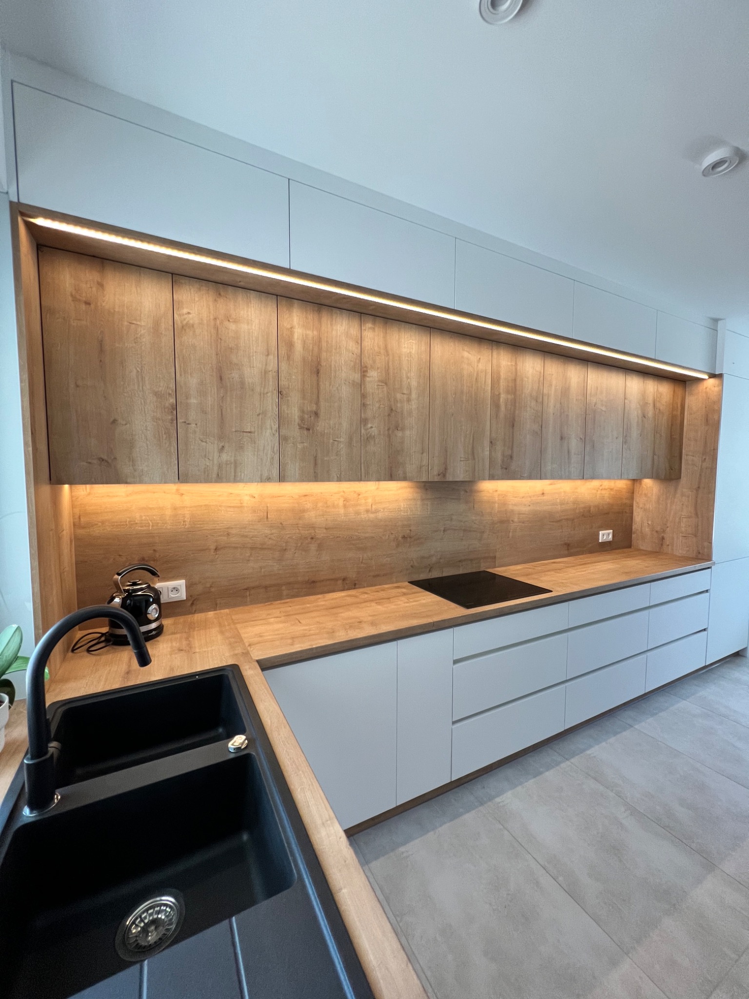 Nowoczesna kuchnia z białymi szafkami dolnymi i górnymi z drewnopodobnym wykończeniem, podświetlona listwą LED, z czarnym zlewem i czajnikiem elektrycznym.