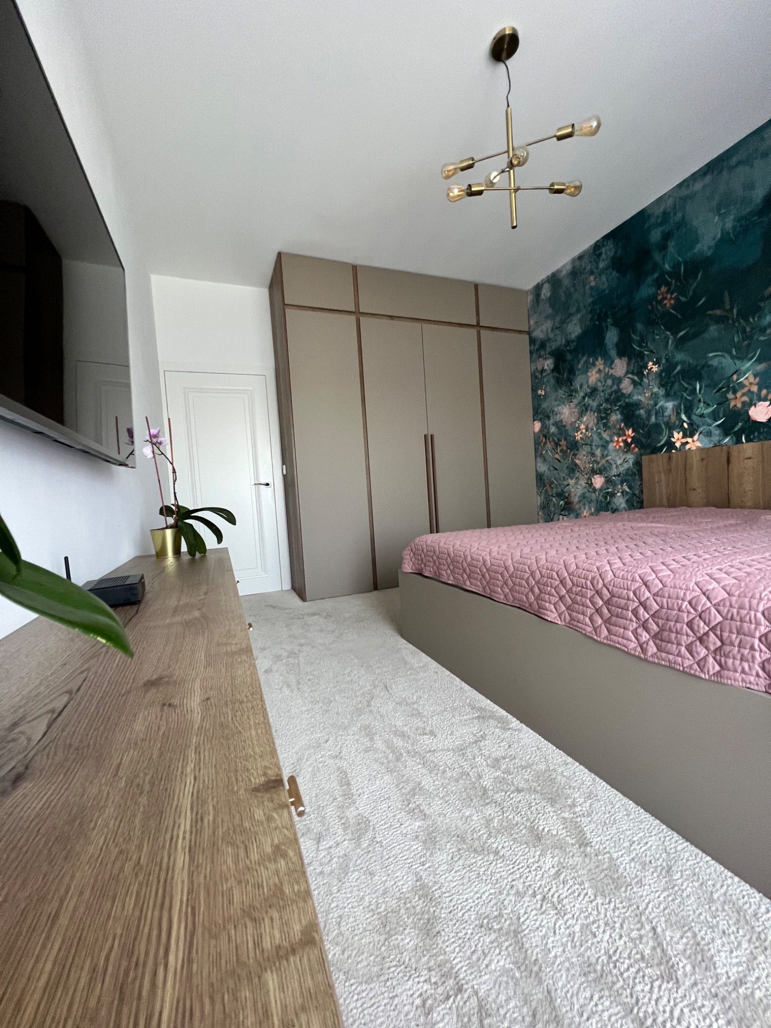 Nowoczesna sypialnia z zabudowaną szafą w odcieniach beżu, łóżkiem z różową narzutą, drewnianą komodą i tapetą w kwiaty na ścianie.