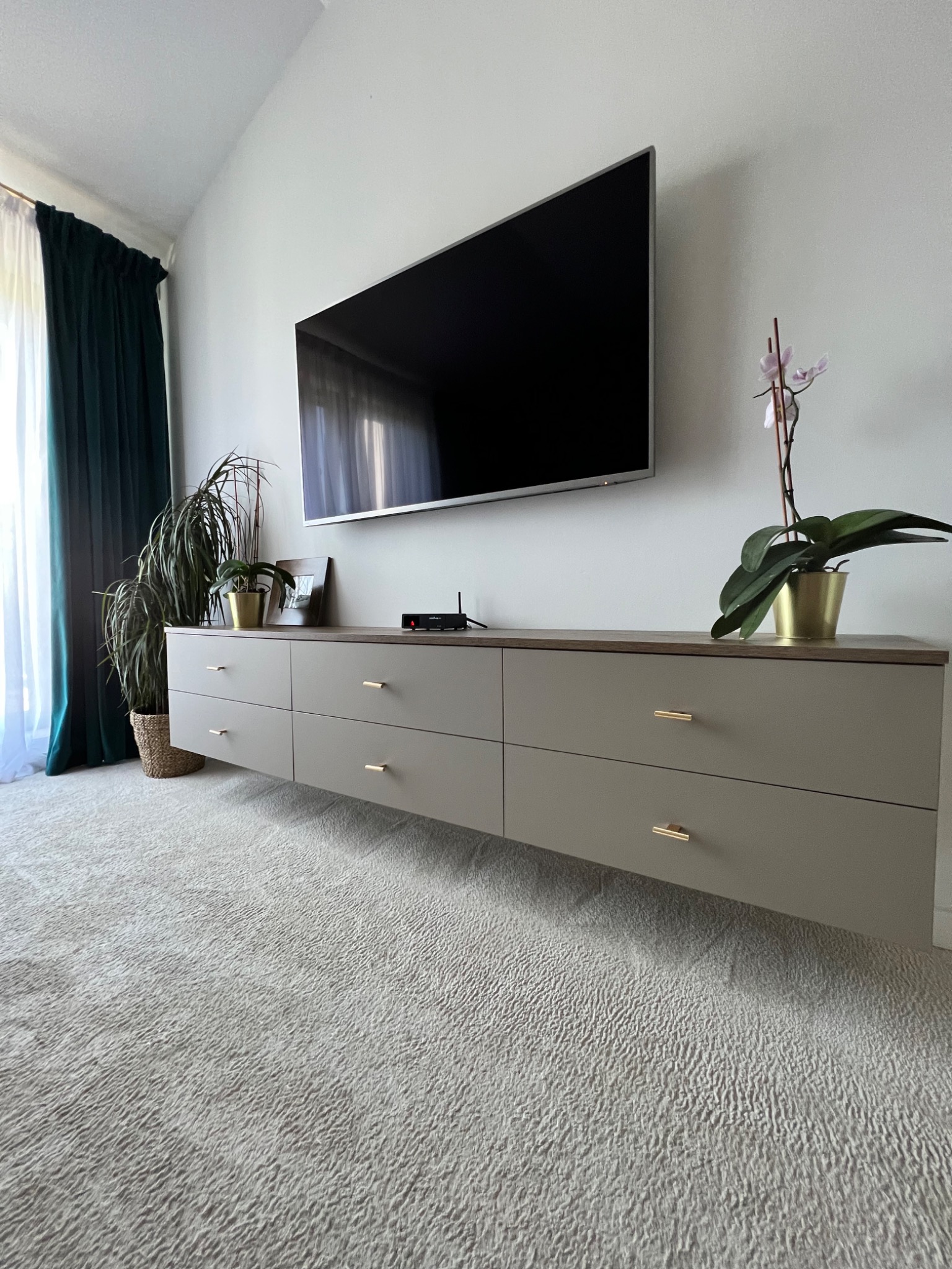 Beżowa szafka RTV z sześcioma szufladami, zawieszona pod telewizorem, z roślinami doniczkowymi na blacie, w nowoczesnym salonie z kremowym dywanem i ciemnozielonymi zasłonami.