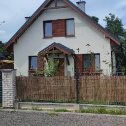 Dwukondygnacyjny dom jednorodzinny z jasną elewacją, ciemnym dachem i drewnianymi elementami wykończeniowymi okien, otoczony płotem z siatki i maty bambusowej, oświetlony girlandą lampek.