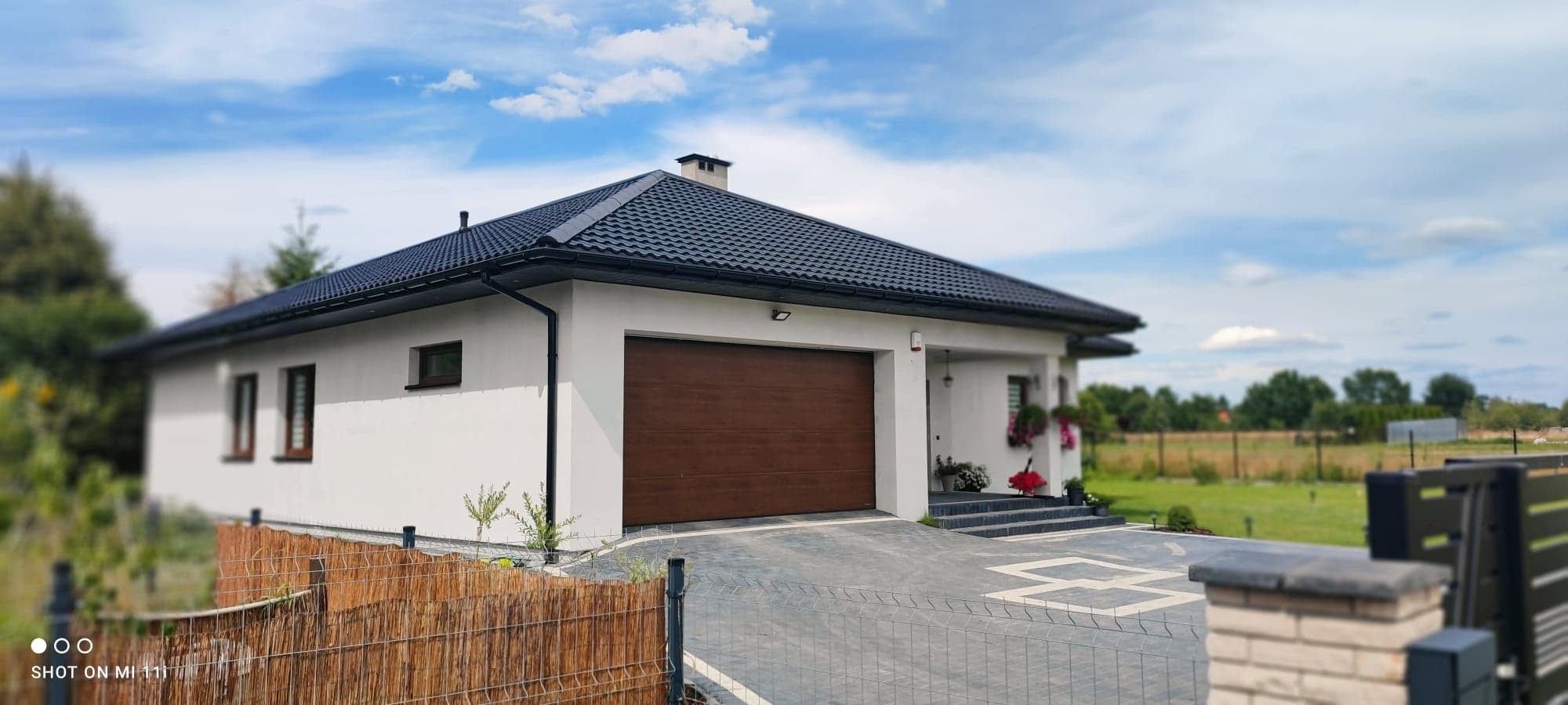 Nowoczesny parterowy dom z brązową bramą garażową i ciemną dachówką, widoczny z perspektywy podjazdu wyłożonego szarą kostką. W tle błękitne niebo z chmurami.