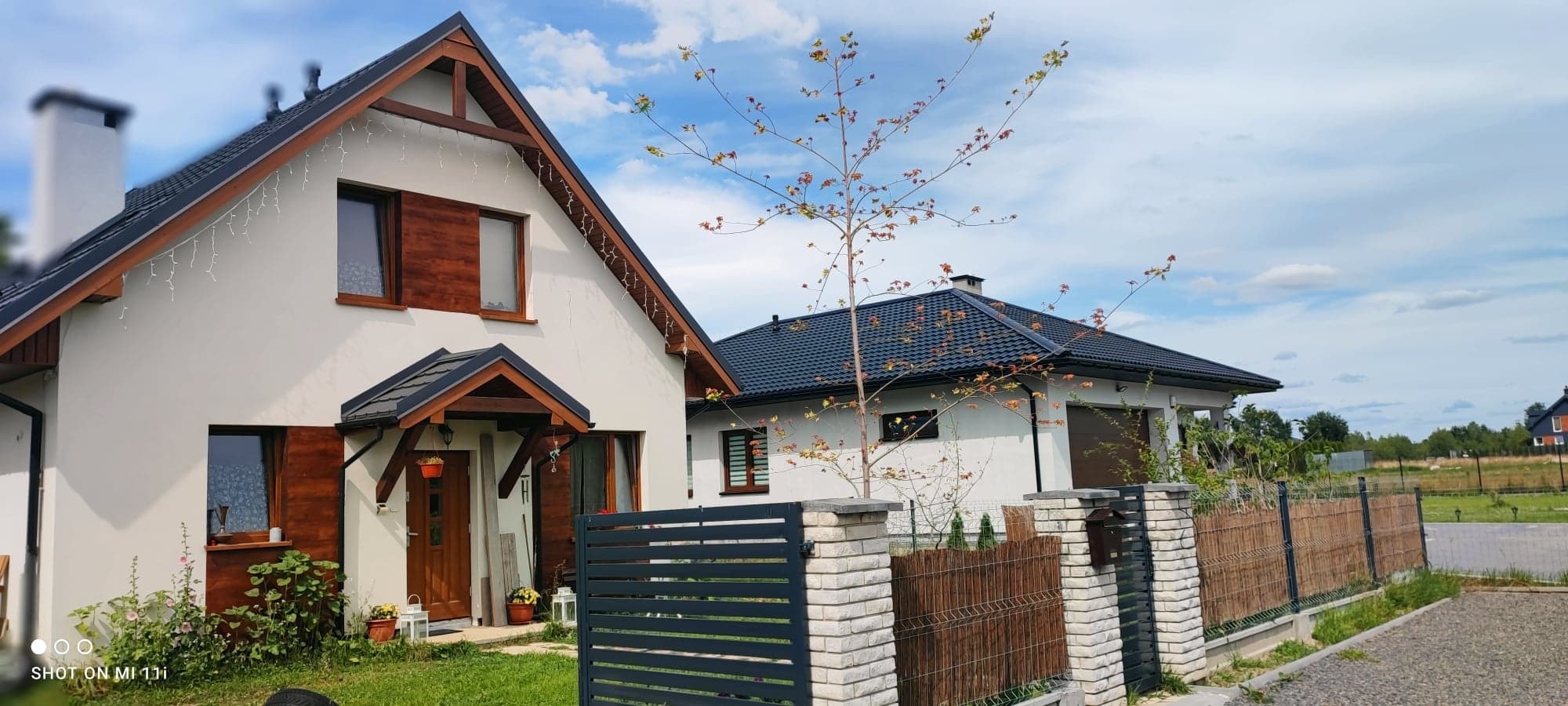 Nowoczesny dom jednorodzinny z białymi ścianami, ciemnym dachem i drewnianymi elementami wykończeniowymi, ogrodzony metalowym płotem na podmurówce z cegły.